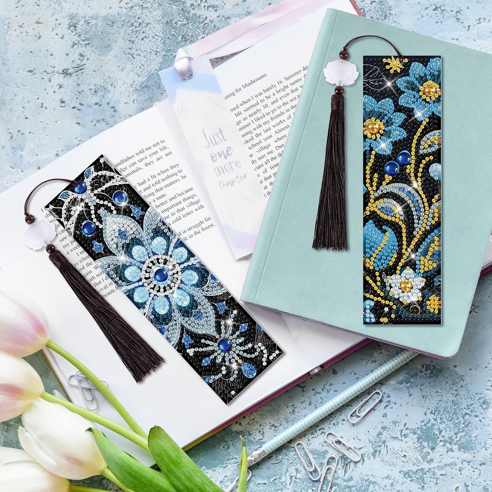Two diamond art bookmarks with floral patterns and tassels are displayed on an open book and a notebook.