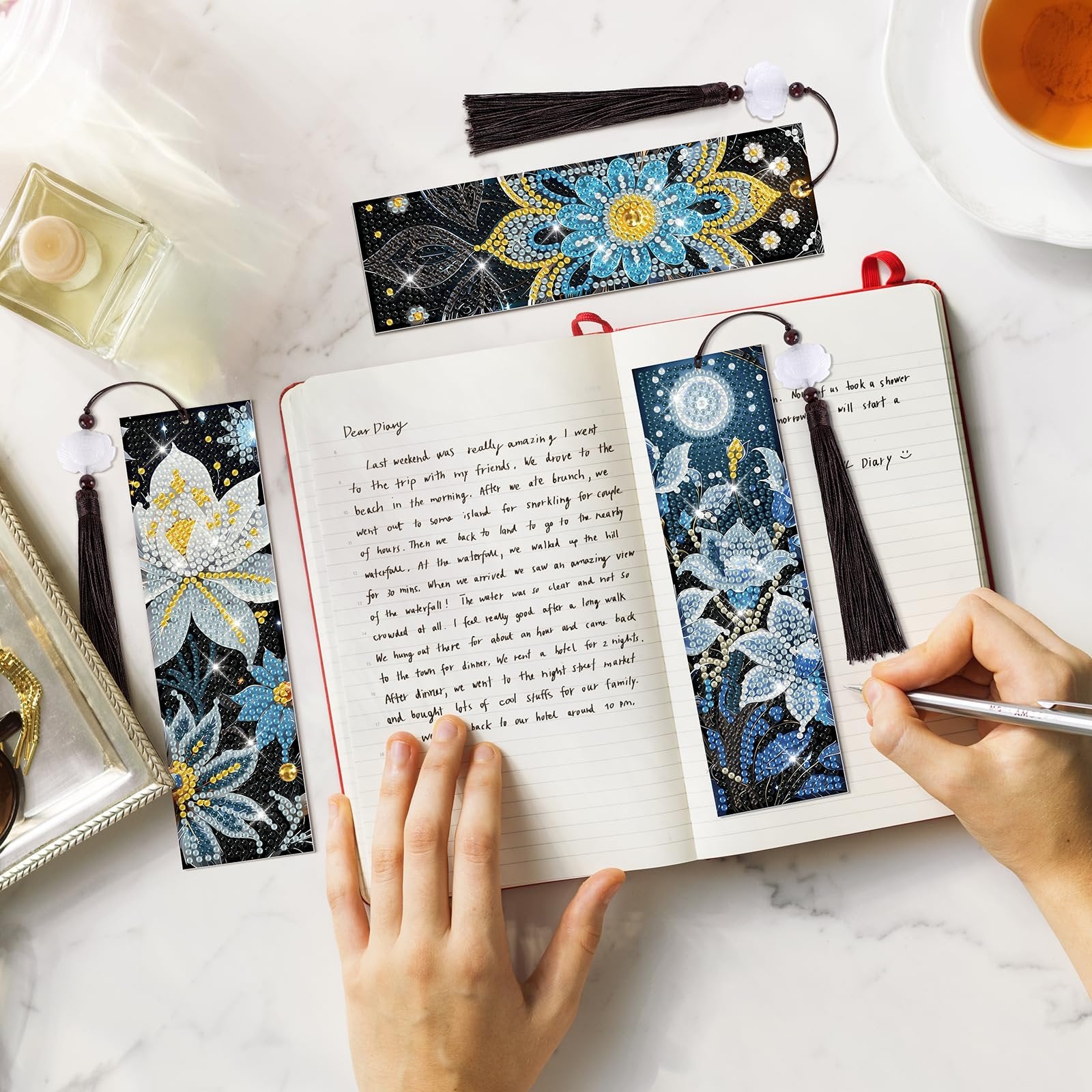 Diamond art bookmarks with tassels laid on an open notebook with handwritten text and a hand holding a pen.