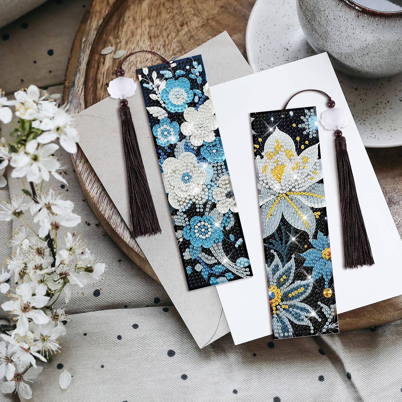Two diamond art bookmarks with floral designs and tassels, displayed on layered paper with a wooden tray and white flowers.