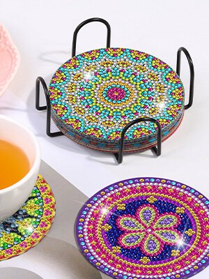 Set of 8 mandala diamond painting coasters with cork bases in a black metal holder, with a cup of tea and another coaster in foreground.