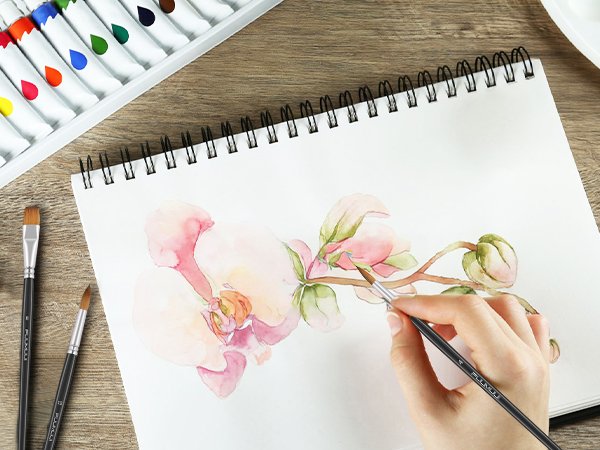 Watercolor painting of a pink orchid with brushes and paint tubes on a wooden desk