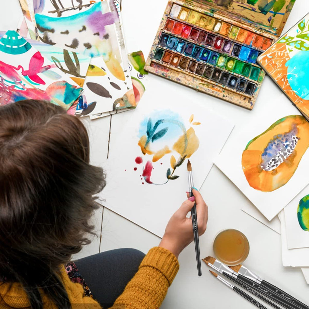 Overhead view of a person painting with a watercolor set and brushes, with artwork and supplies spread out on a white surface.