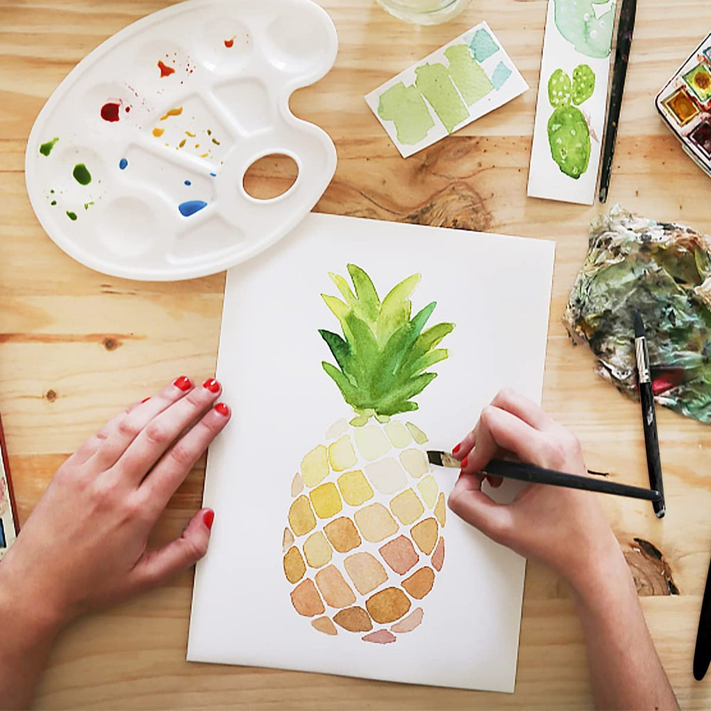 Hands painting a watercolor pineapple on white paper with a palette and brushes on a wooden table