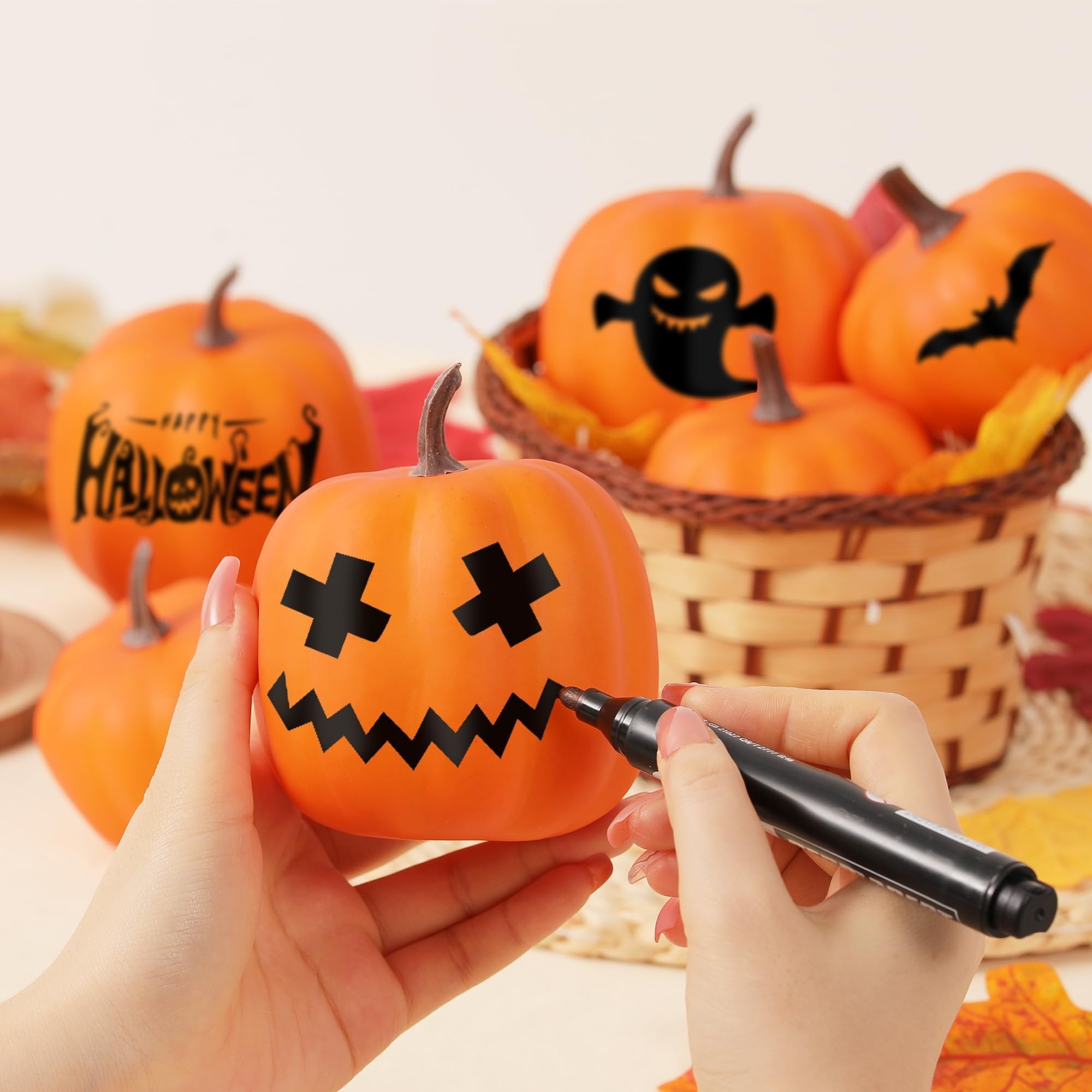 Hands decorating an orange artificial pumpkin with a black marker for Halloween, with other pumpkins in a basket.