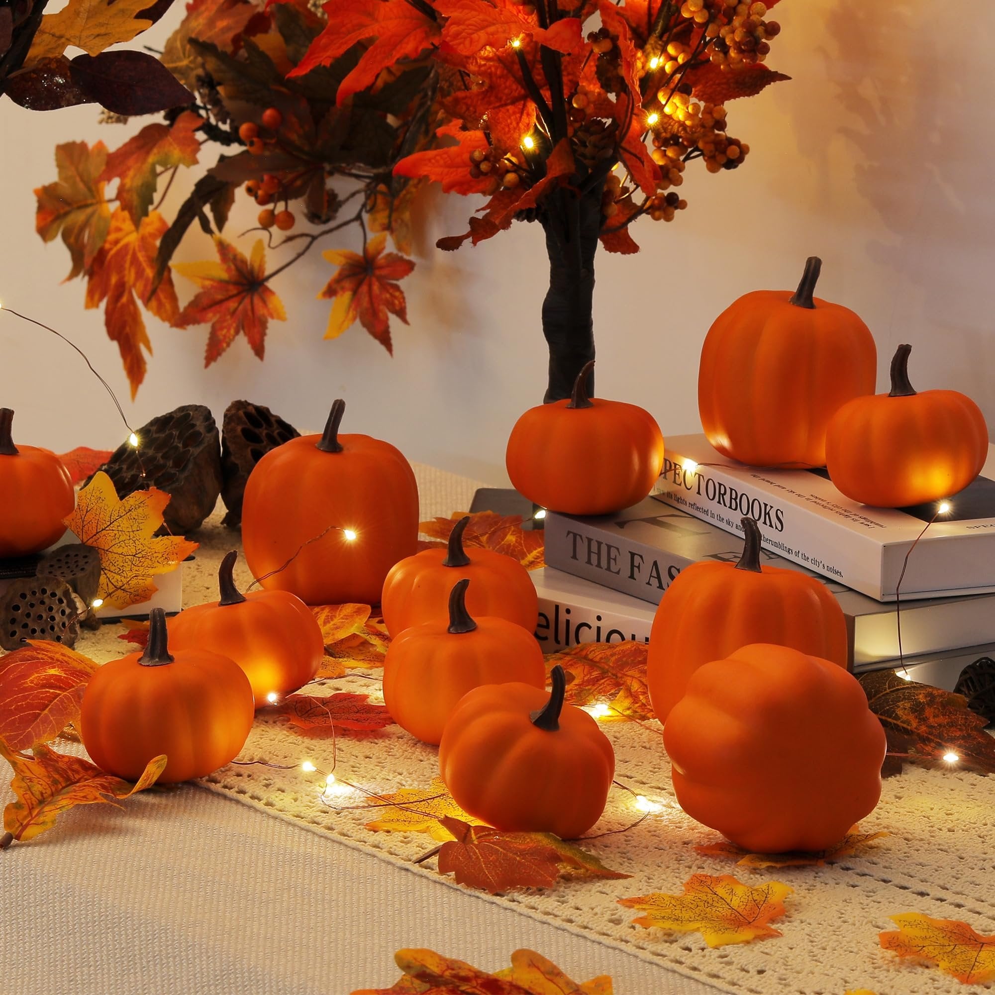 Set of artificial orange pumpkins arranged with fall leaves and string lights on a textured surface, with books in the background.