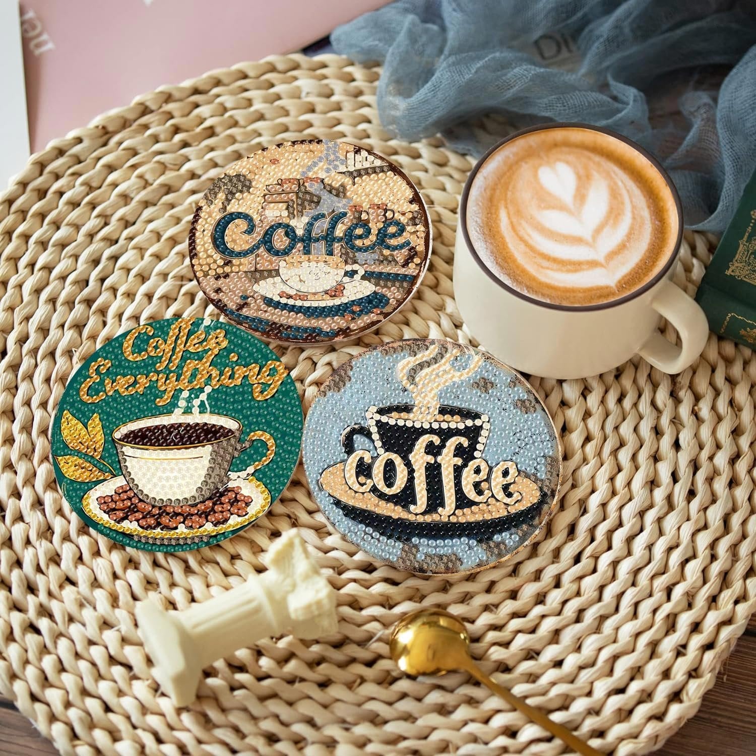 Three diamond art coasters with coffee designs and a latte art coffee cup on a woven placemat.