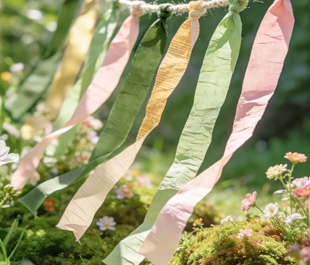 Crepe paper streamers in pastel green, sage, yellow, and pink hanging on a twine string outdoors with flowers and greenery.