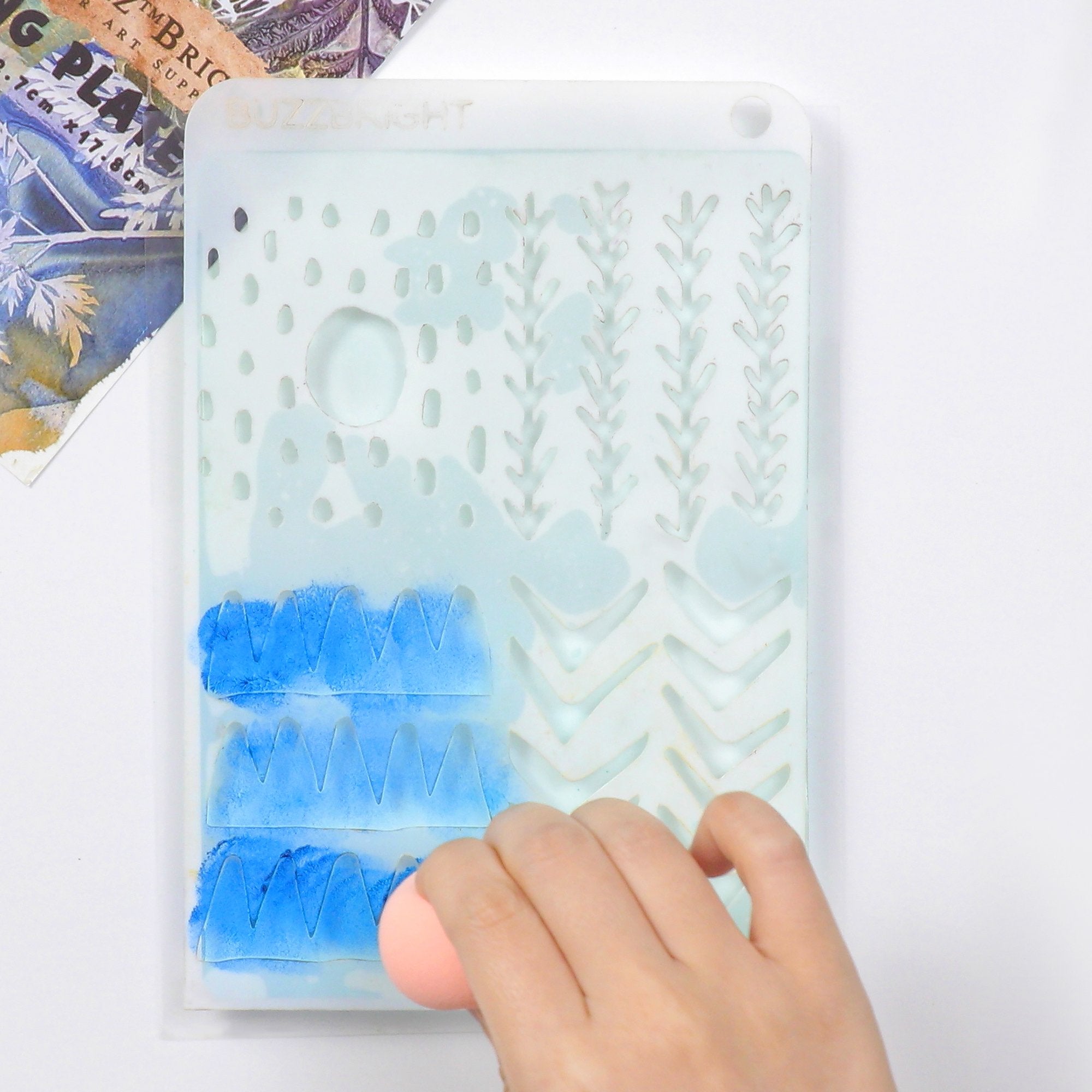 Gel printing plate with stencil patterns being applied with blue paint and a sponge by a hand.