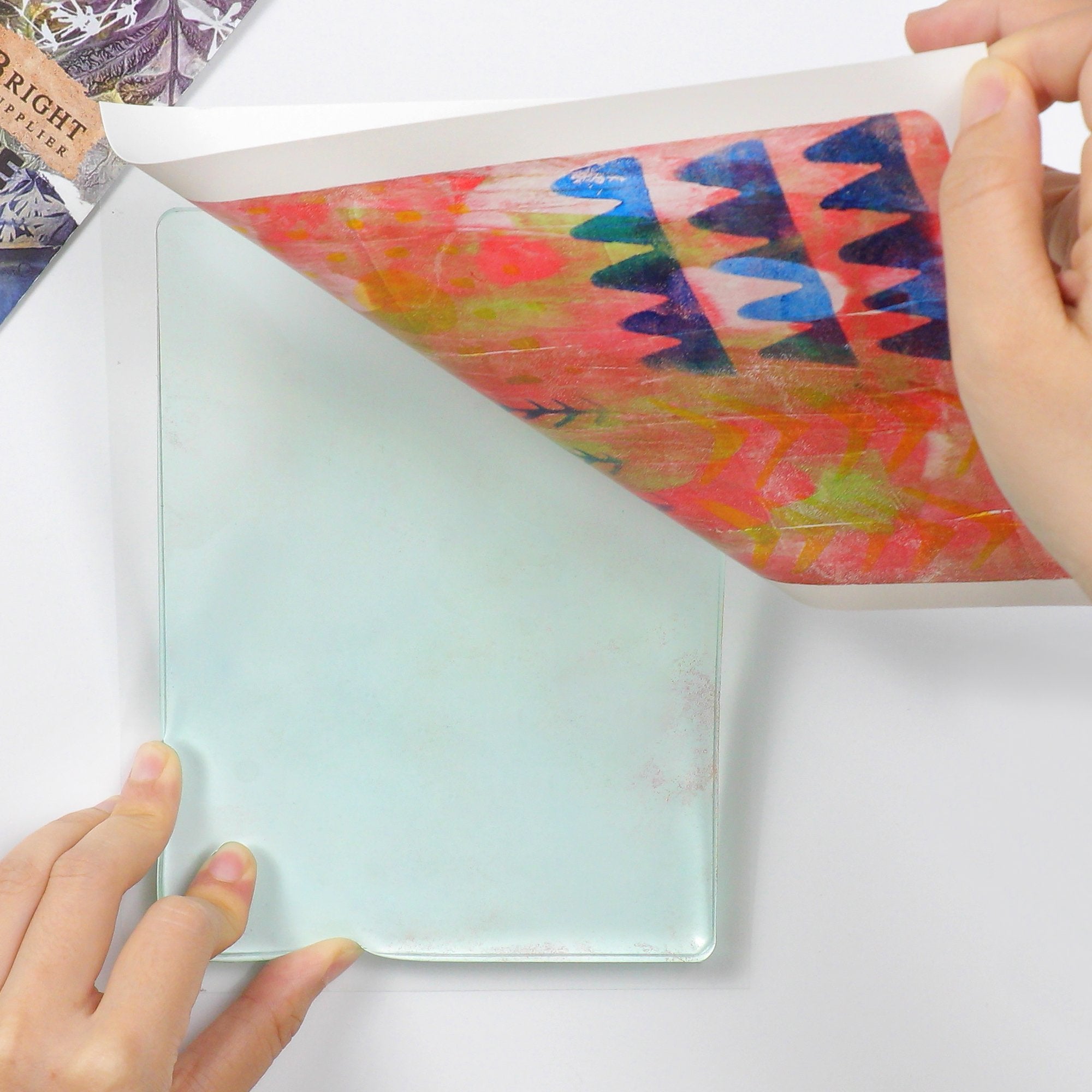 Hands peeling a colorful abstract print from a light blue gel printing plate on a white surface.