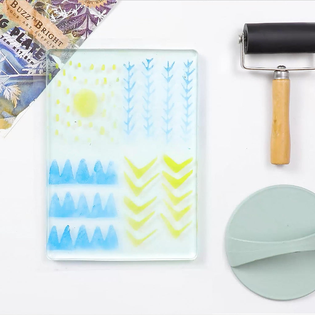 Gel printing plate with blue and yellow paint designs, brayer, and green scraper tool on a white surface.