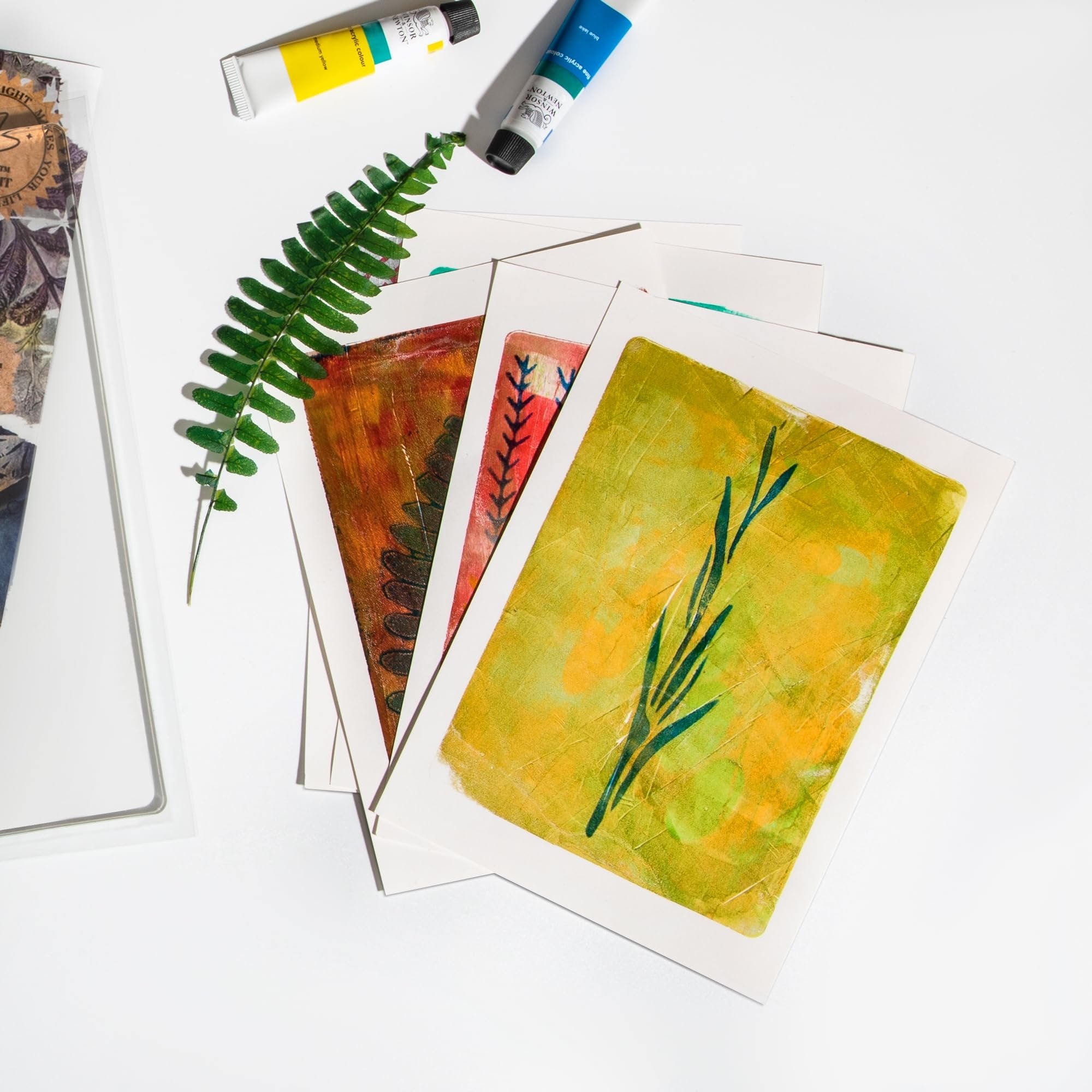 Gel prints with botanical designs and acrylic paint tubes on a white surface