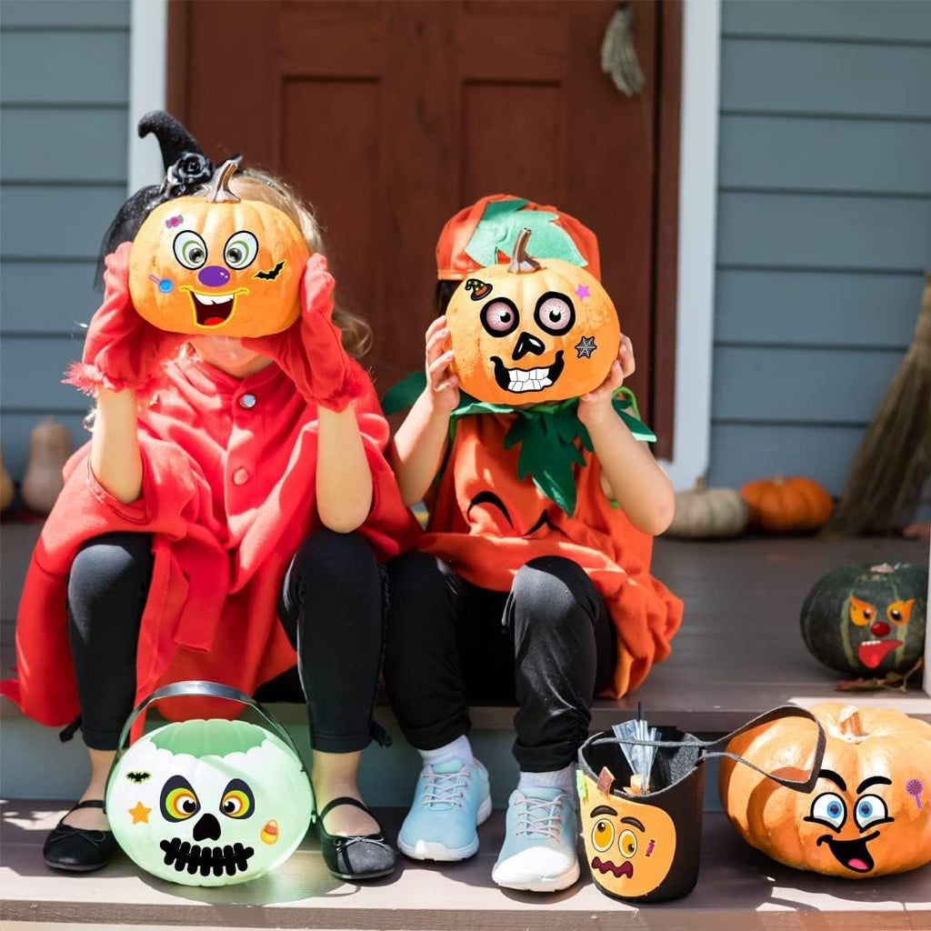 Two children in Halloween costumes sit on a porch holding decorated pumpkins; candy buckets are in front.