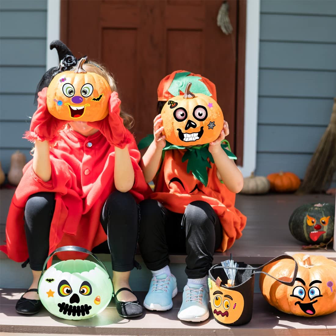 Two children in Halloween costumes sit on porch steps holding decorated pumpkins; candy buckets and pumpkins are on the steps.