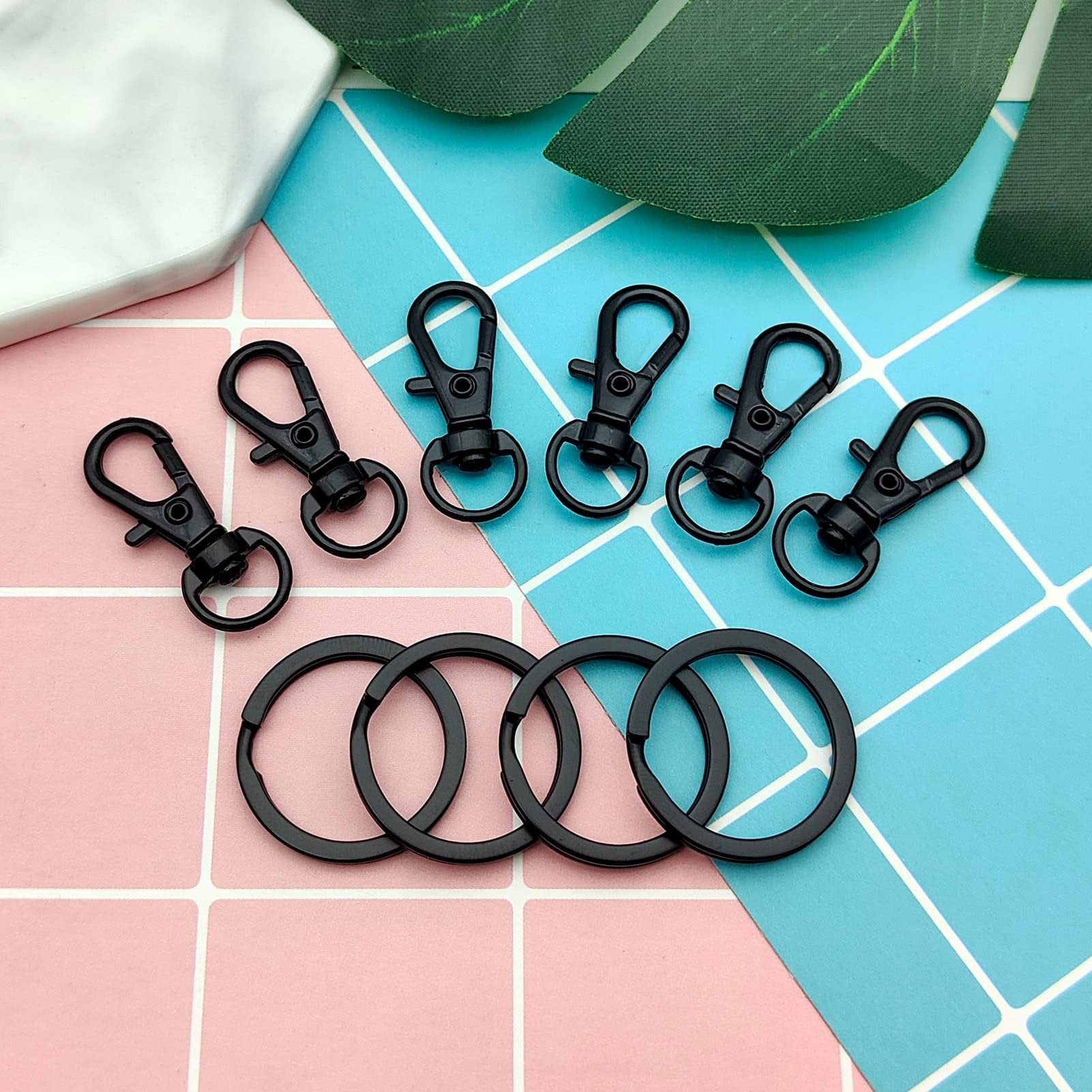 Black swivel snap hooks and key rings arranged on a pink and blue grid background with decorative leaves.