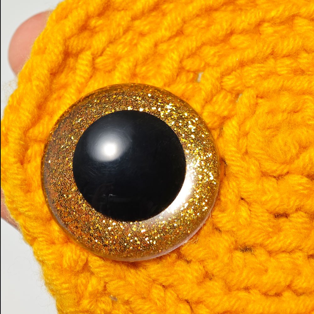 Close-up of a large, glittery gold safety eye with a black pupil, resting on bright orange yarn.