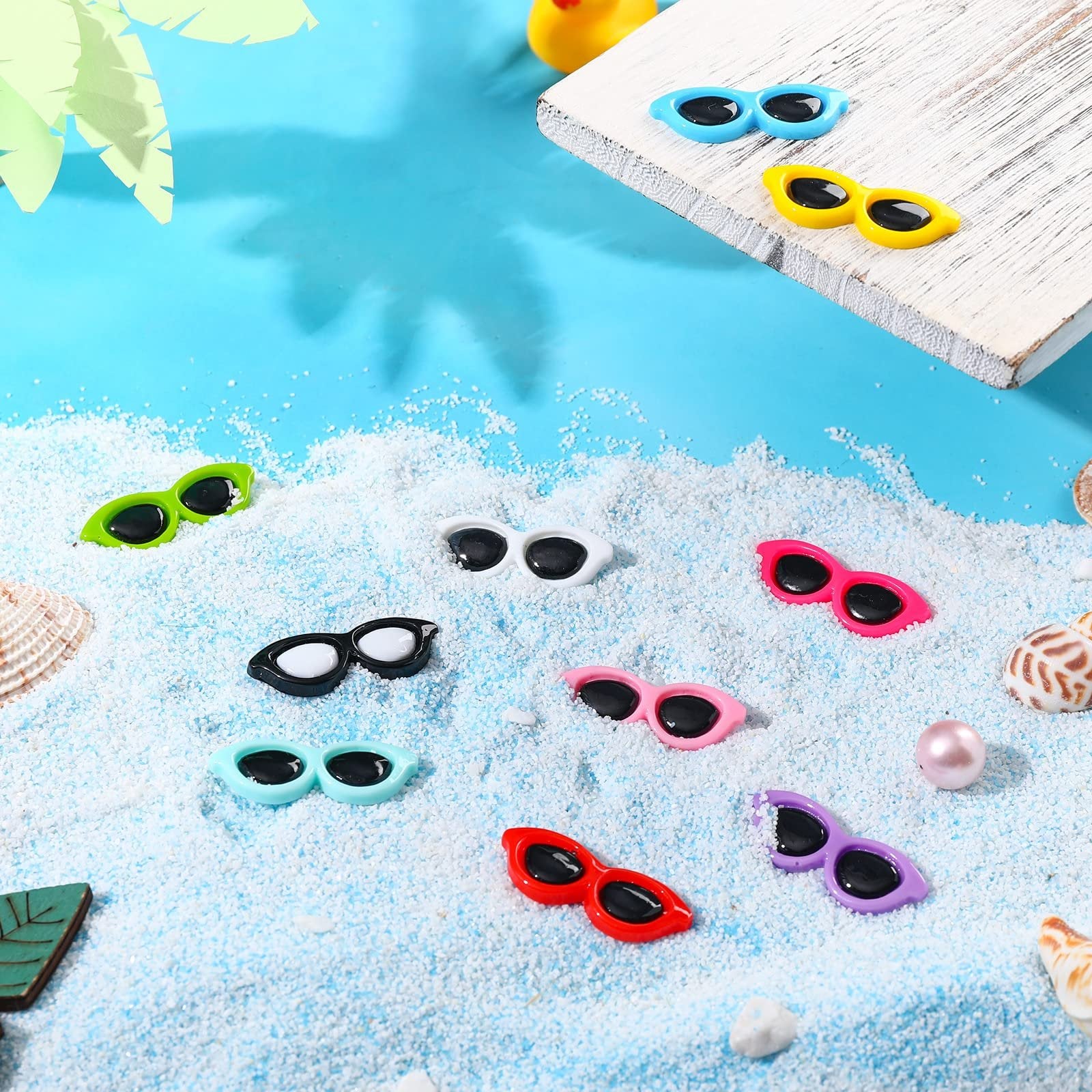 Assorted colorful mini sunglasses resin flatback charms scattered on white sand with seashells and palm leaf shadows.