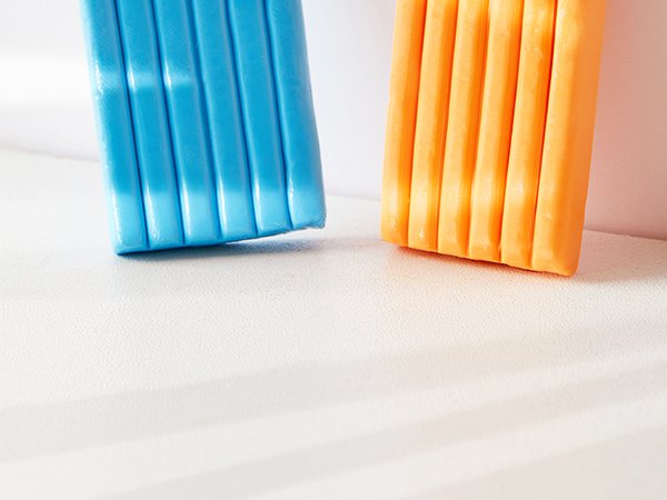 Two bars of polymer clay, one blue and one orange, shown from a low angle on a textured white surface with soft shadows.
