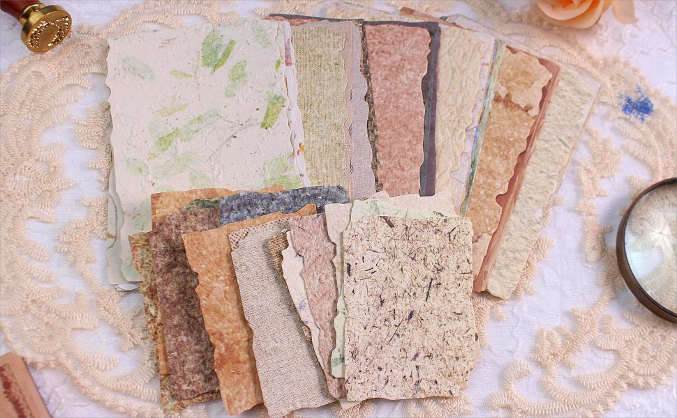 Assortment of textured, deckled-edge scrapbook papers with vintage collage designs and embedded natural elements, arranged on lace.