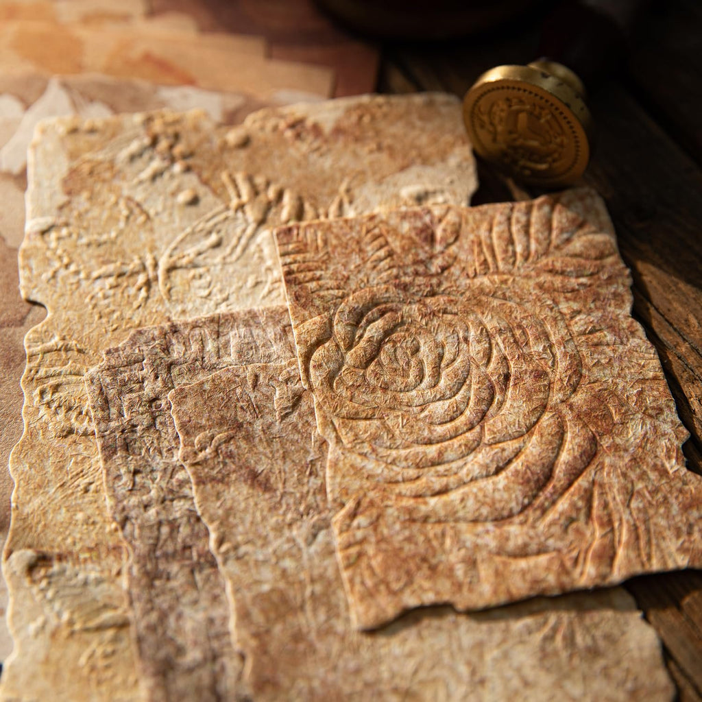 Close-up of textured, embossed scrapbook paper with deckled edges and vintage floral designs, next to a gold wax seal stamp.