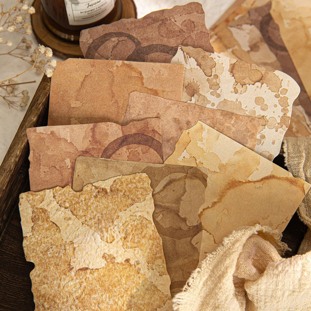 Textured scrapbook paper with vintage coffee stain designs and deckled edges, arranged in a wooden tray with burlap.