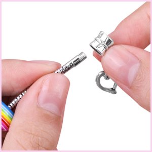 Hands assembling a silver snake chain bracelet with a heart-shaped clasp and rainbow-colored beads.