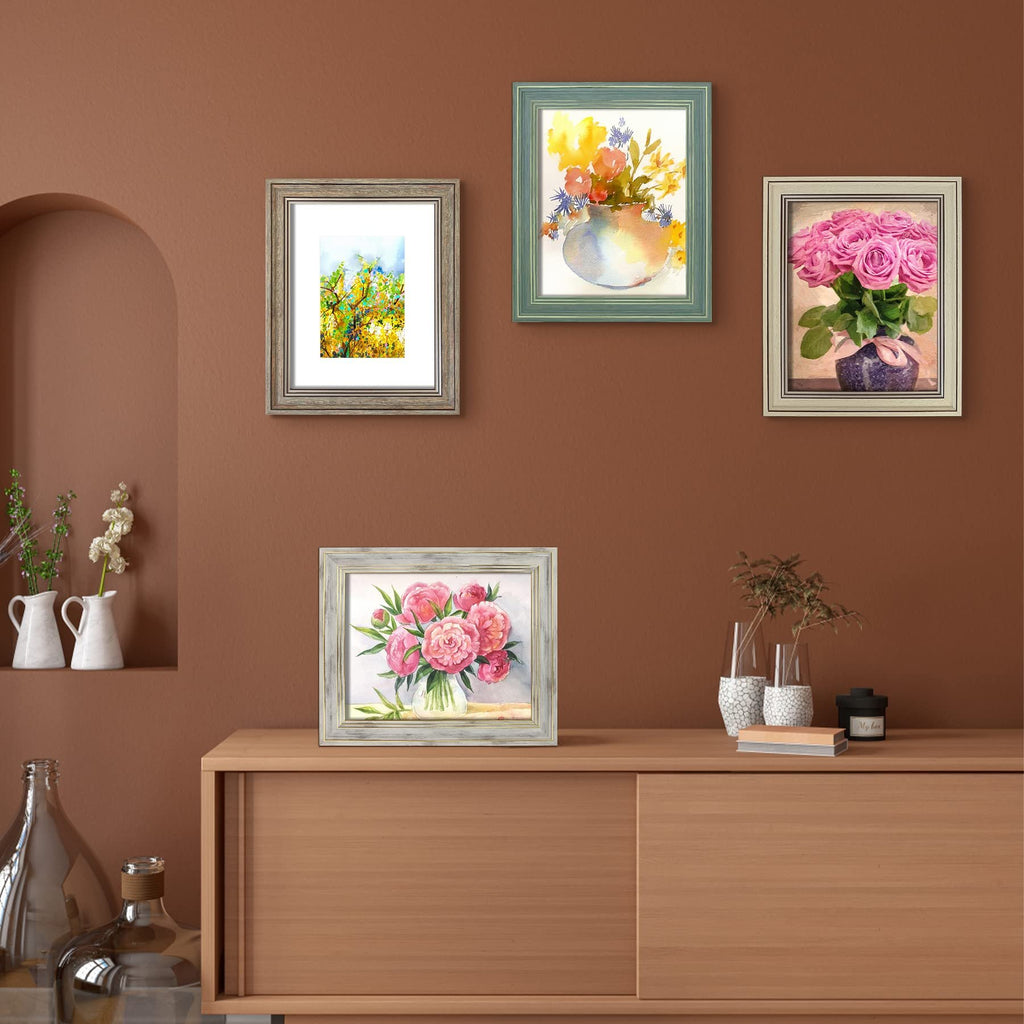 Four framed watercolor paintings displayed on a brown wall above a wooden cabinet. One frame is on the cabinet, showing pink peonies. Three frames hang on the wall: one with yellow and green abstract art, one with a vase of flowers, and one with pink roses.