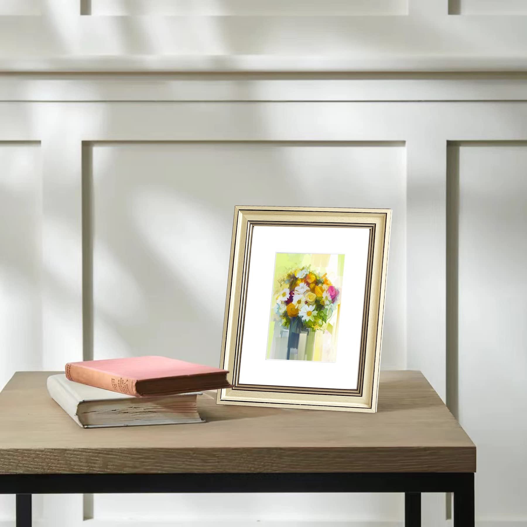 Gold picture frame with floral art on a wooden table with two books, against a white paneled wall.