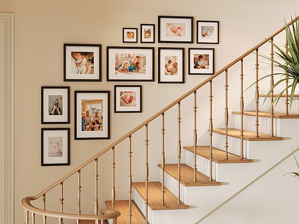 Gallery wall of eight black picture frames with family photos displayed on a wall next to a wooden staircase.