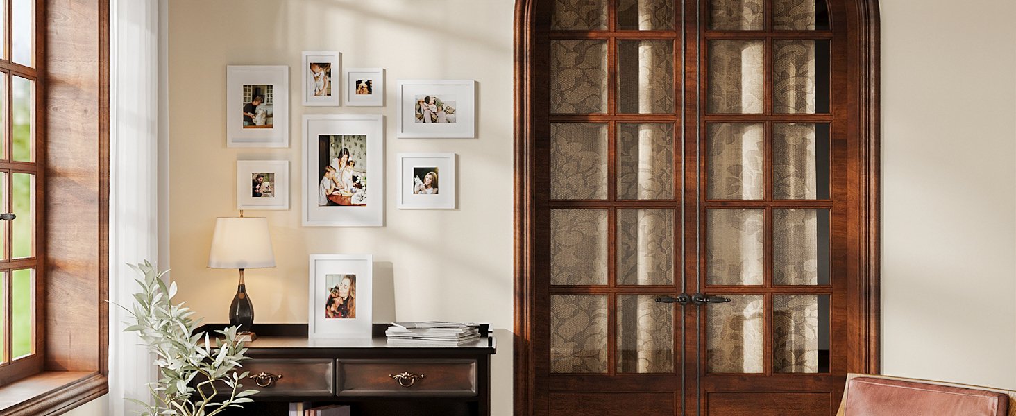 Gallery wall of white picture frames on a wall above a dark wood desk with a lamp and plant.