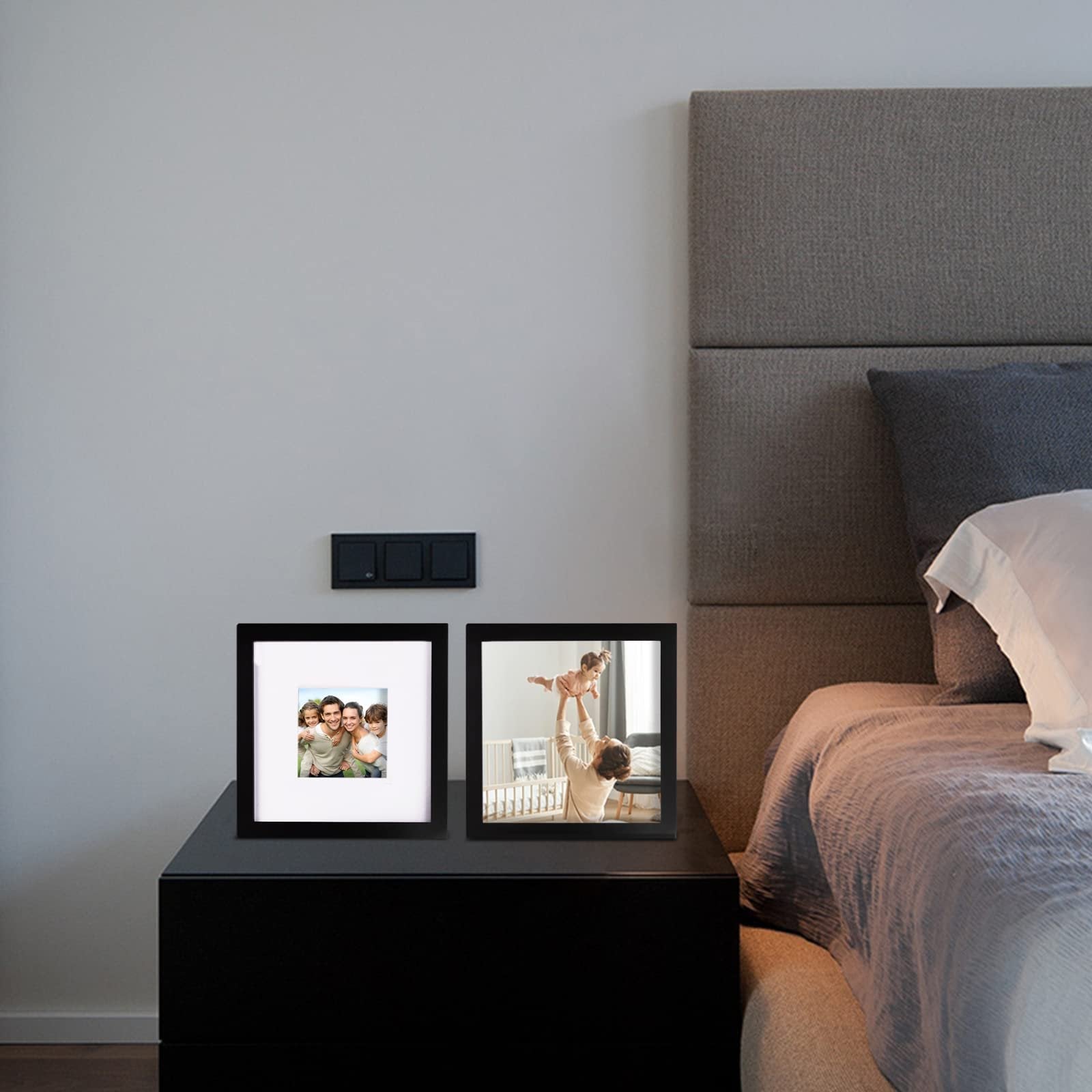 Two black picture frames on a nightstand next to a bed, one showing a family, the other a parent with a baby.