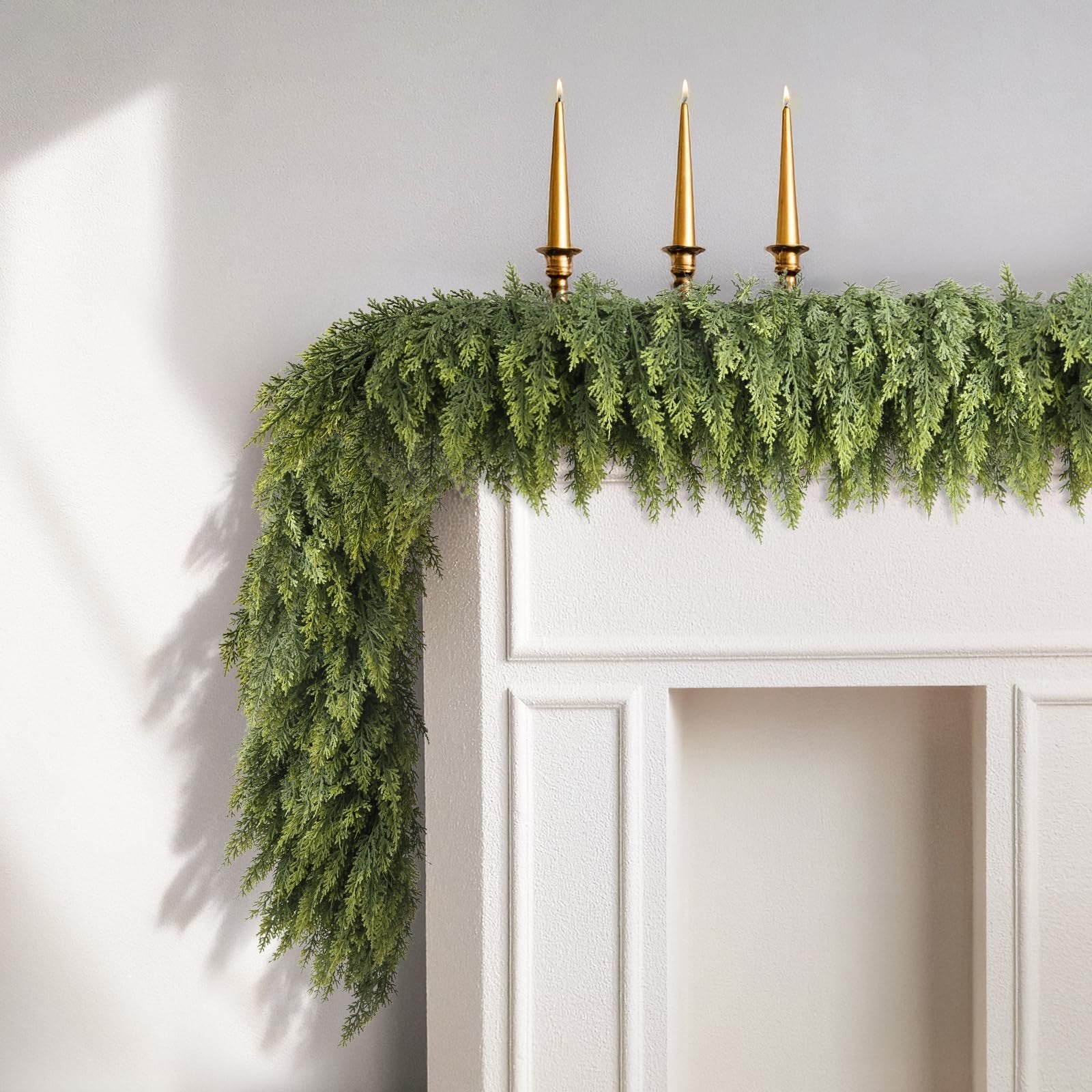 Artificial green cedar garland draped over a white mantelpiece with three gold candlesticks