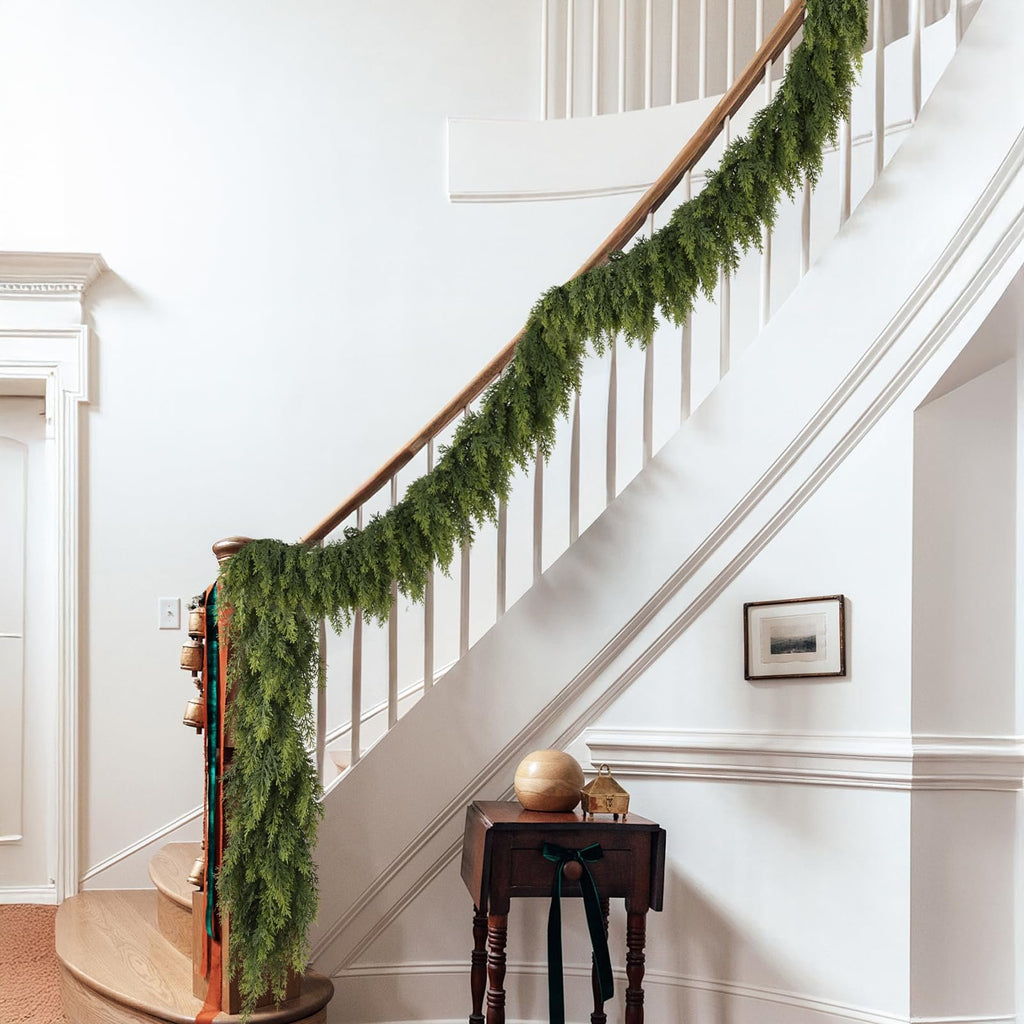 Green artificial cedar garland draped along a wooden banister on a staircase, with decorative bells and ribbons on the newel post.