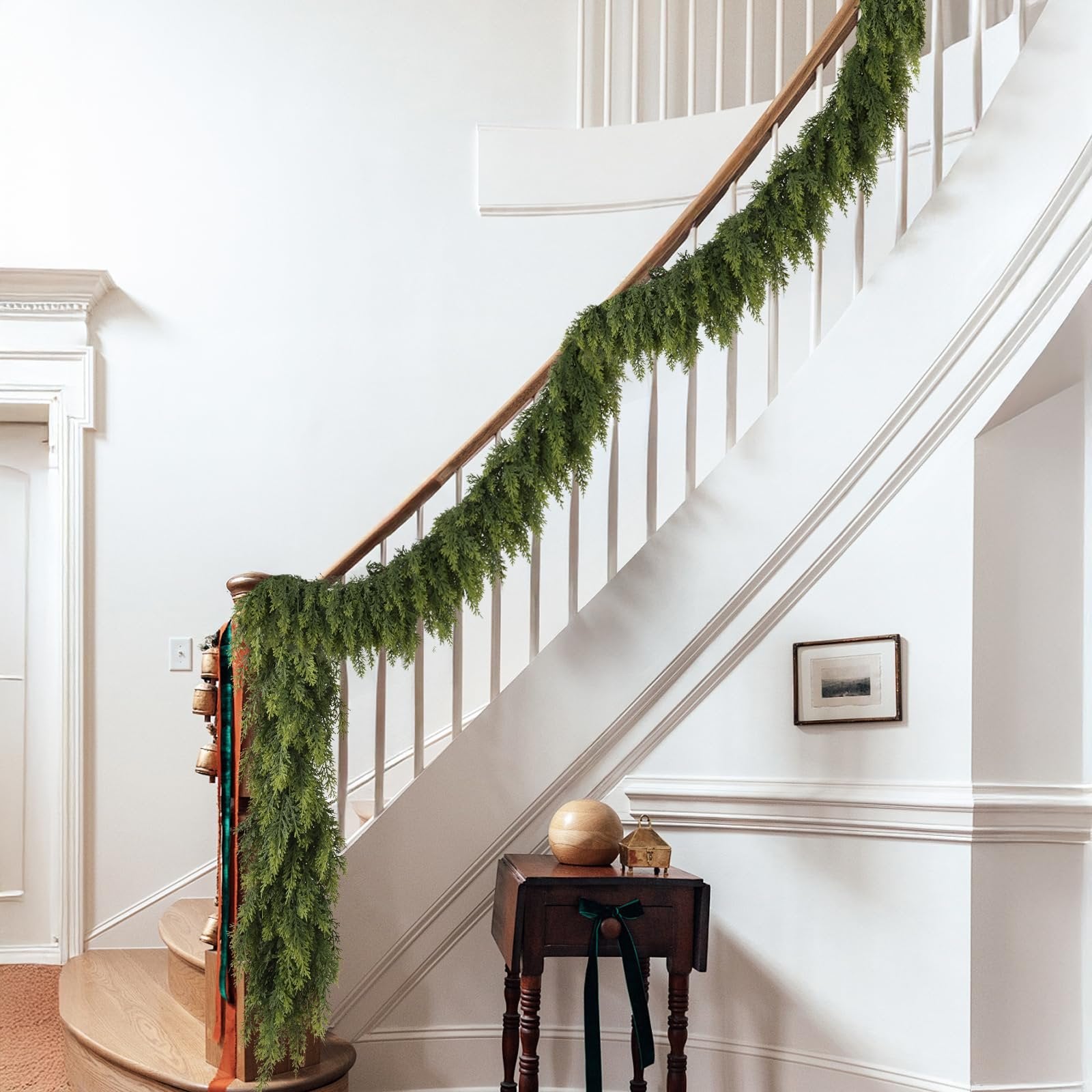 Green artificial cedar garland draped along a wooden banister on a staircase, with decorative bells and ribbons on the newel post.