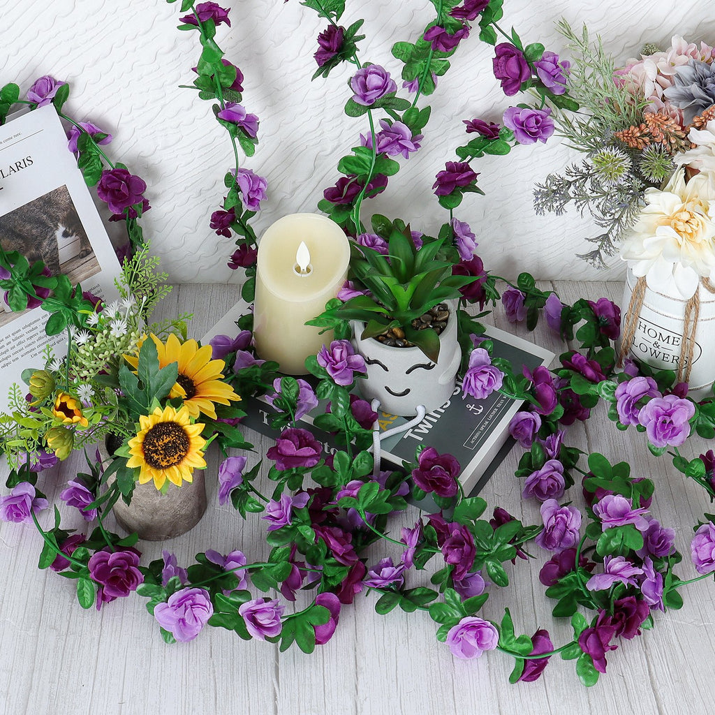 Artificial purple rose garland draped around decorative items including a candle, potted plants, and books.