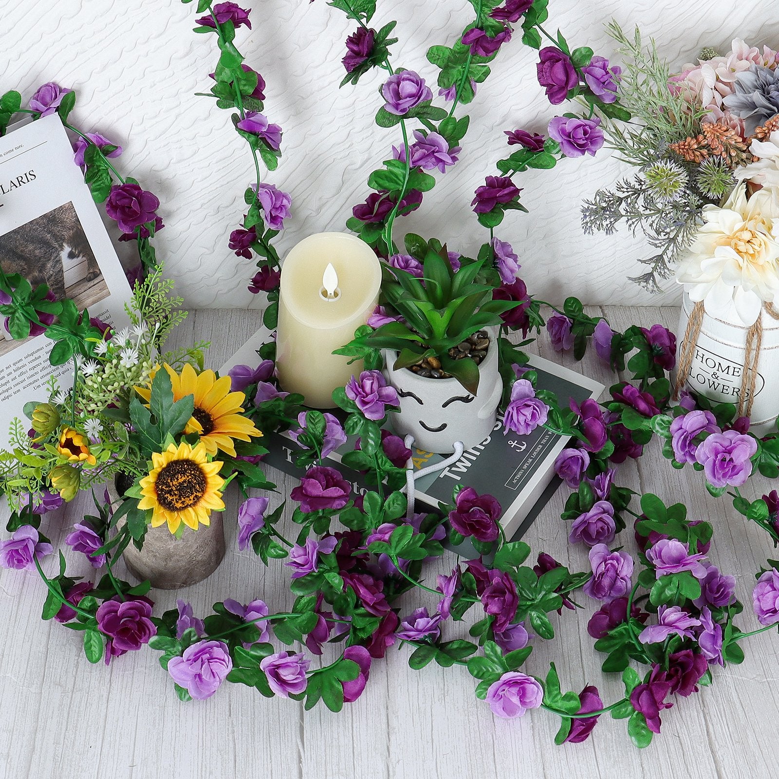 Artificial purple rose garland draped around decorative items including a candle, potted plants, and books.