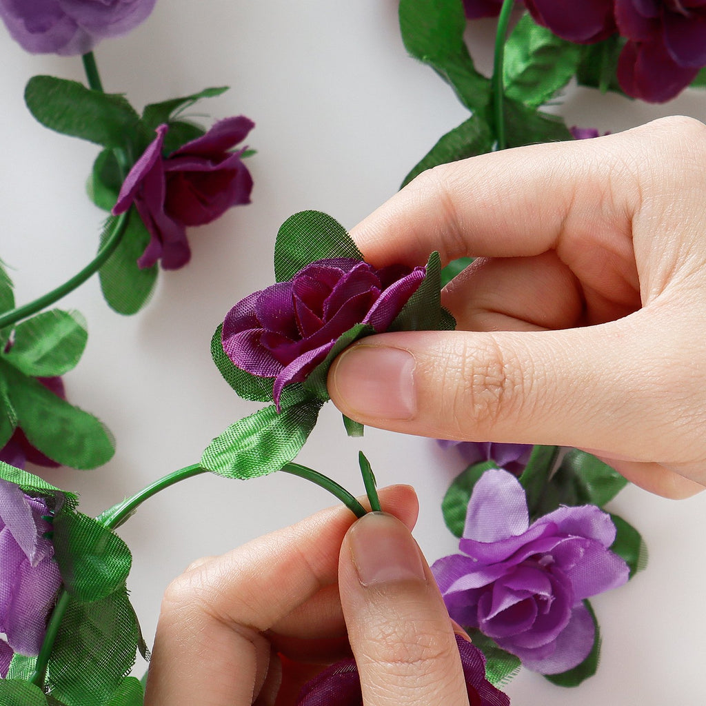 Hands arranging artificial purple rose garland with green leaves on a white background.