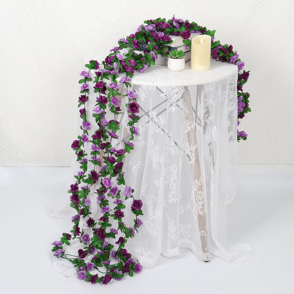 Artificial purple rose garland draped over a white lace-covered table with books and a candle.