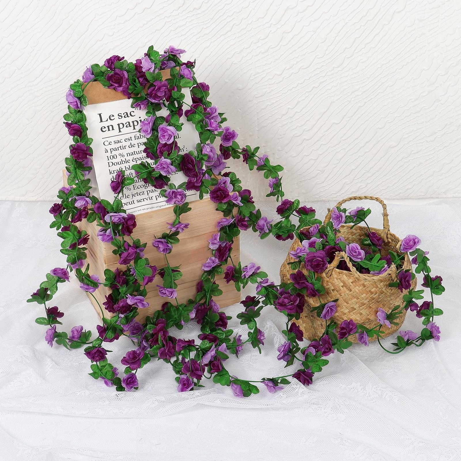 Artificial purple rose garland draped over a wooden box and spilling into a woven basket, with a paper bag visible.