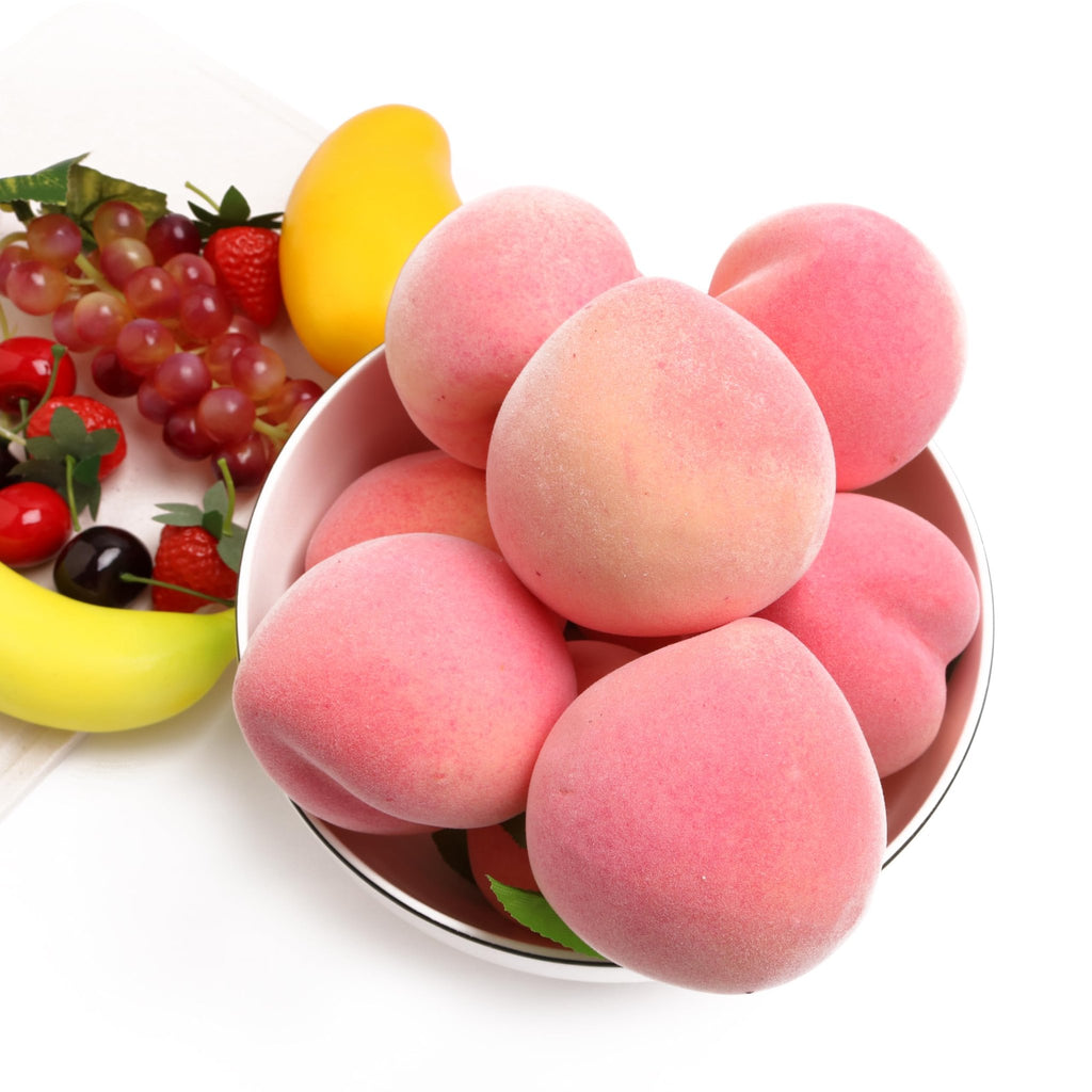 Artificial pink peaches in a bowl with grapes, strawberries, a banana, and a mango on a white surface.