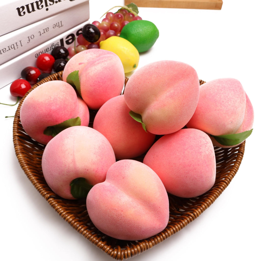 Pile of artificial pink peaches in a woven basket with grapes, lemon, lime, and cherries in the background.