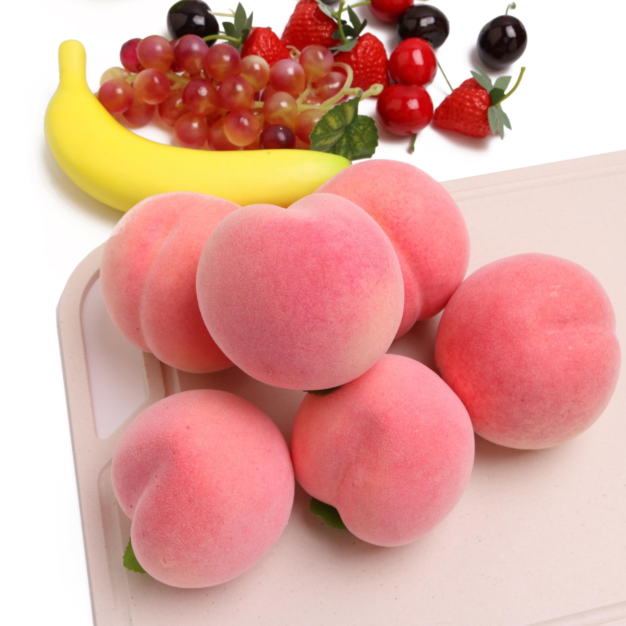 Artificial pink peaches and assorted fake fruits including grapes, strawberries, cherries, and a banana on a white background.