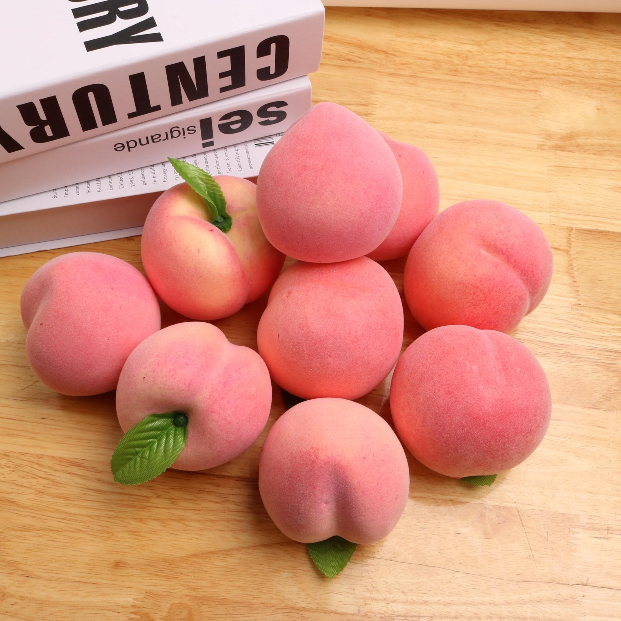 Nine artificial pink peaches with green leaves scattered on a wooden surface, with books in the background.