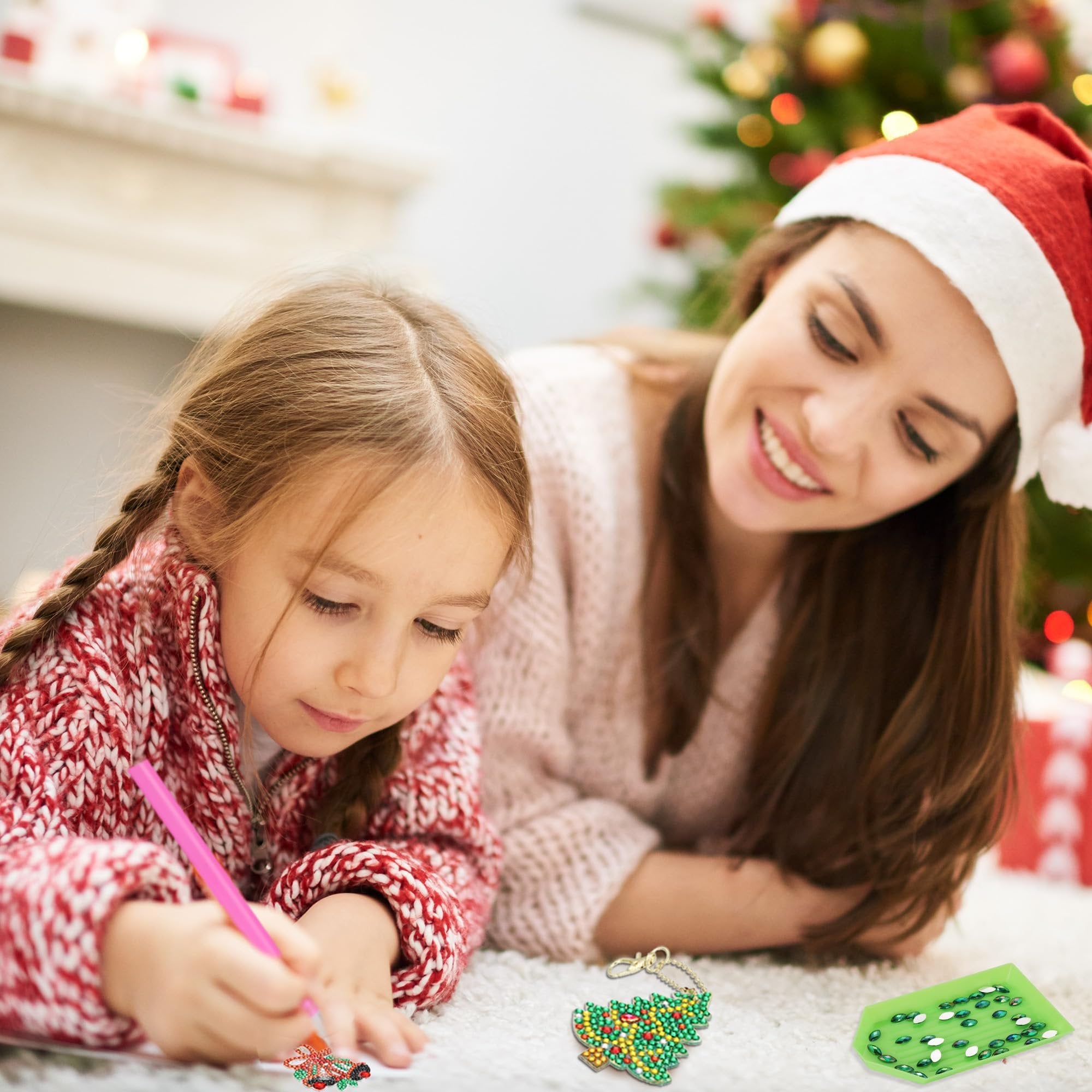 Mother and daughter doing diamond art Christmas ornaments on a rug near a fireplace and tree.