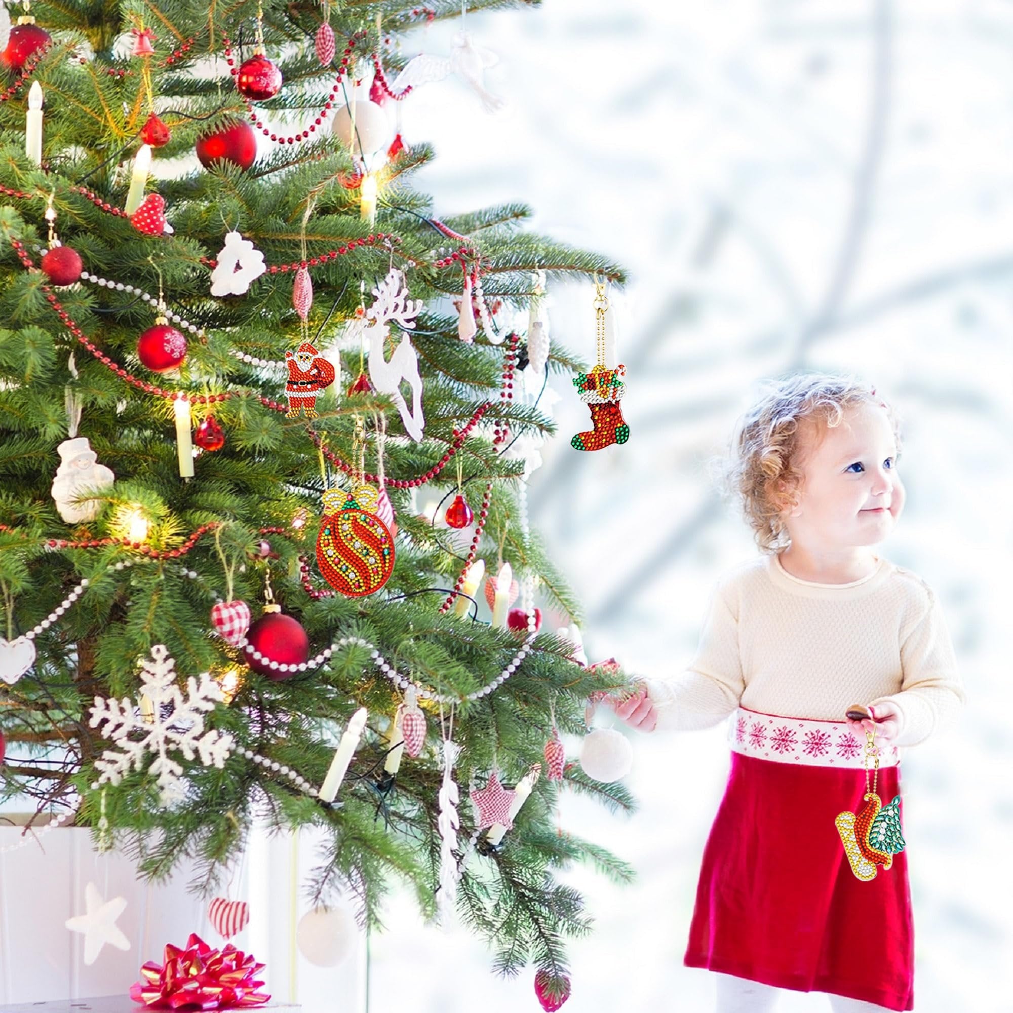 Toddler girl holding diamond art Christmas ornament keychains near a decorated tree