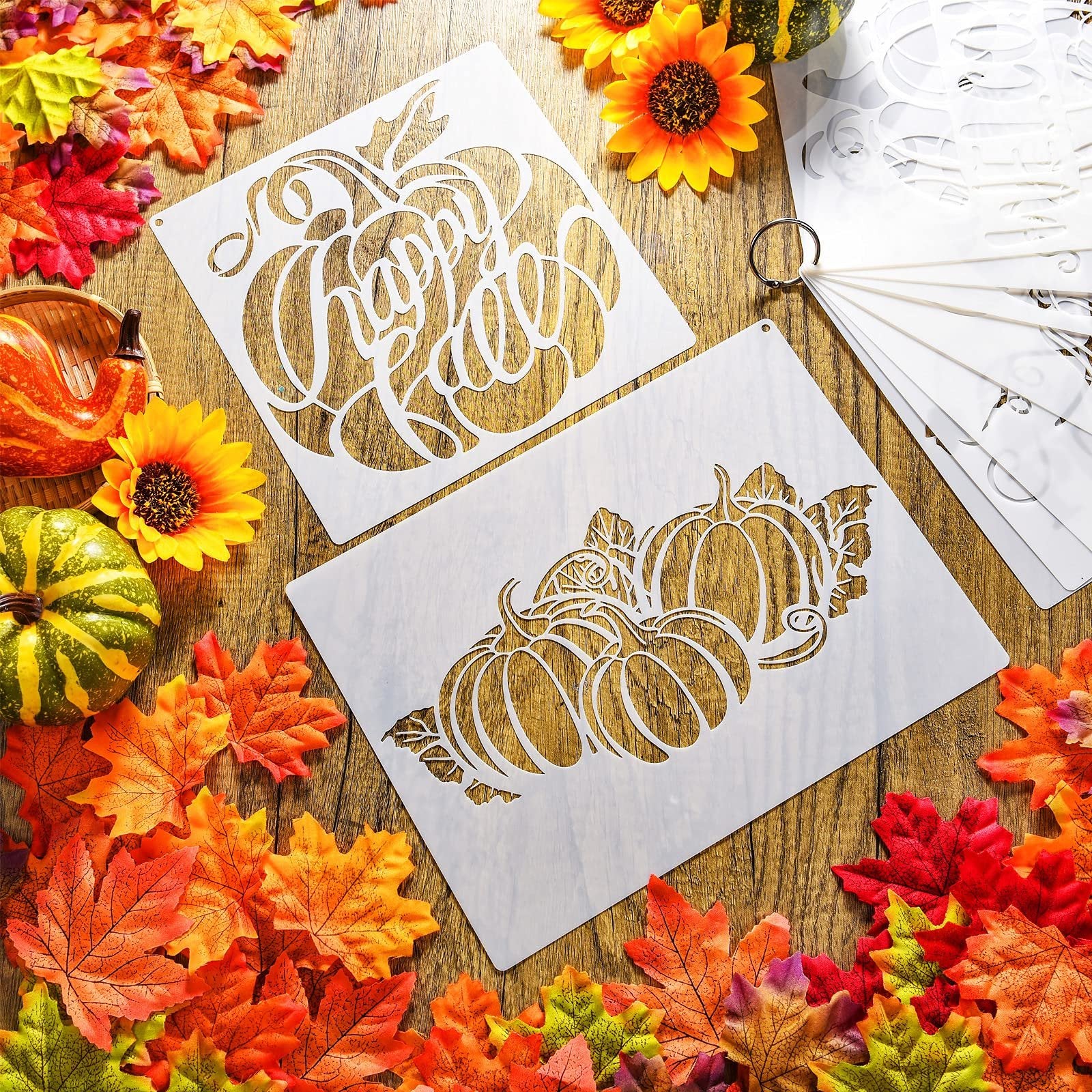 Two fall pumpkin stencils with 'Happy Fall' text and a pumpkin cluster design, surrounded by autumn leaves and decorative gourds on a wooden surface.