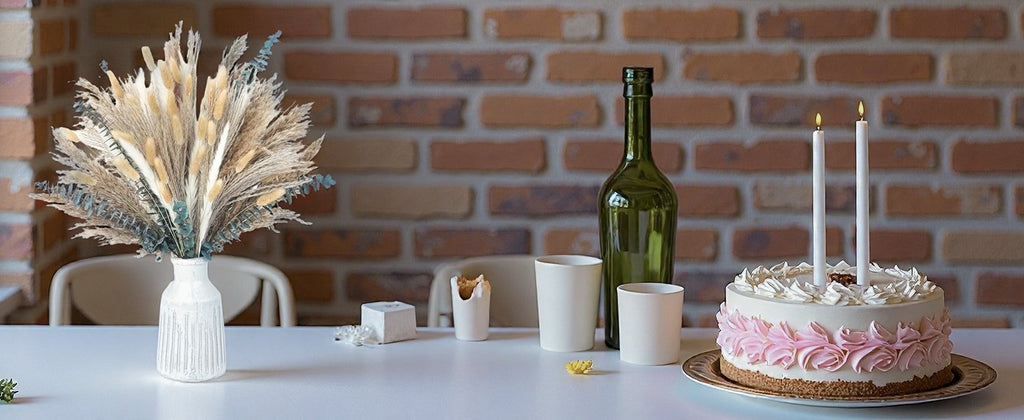Dried flower arrangement in a vase next to a cake with candles and a bottle on a table