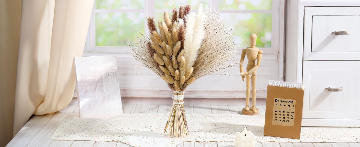 Bouquet of dried flowers including bunny tails and pampas grass, arranged with stems tied together, displayed on a table with decorative items.