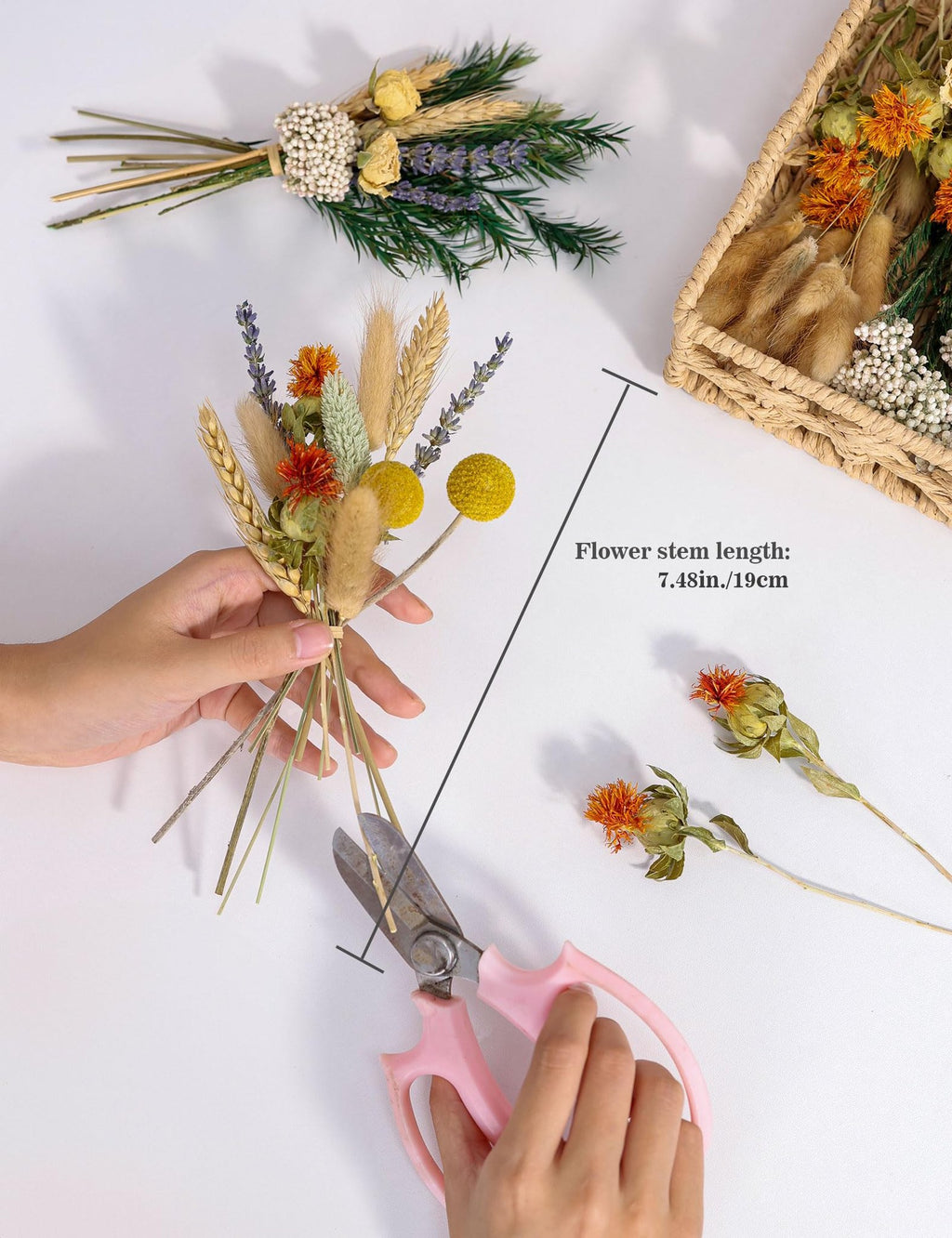 Hands holding a bouquet of dried flowers with wheat and lavender, pink scissors nearby, and a woven basket with more flowers.