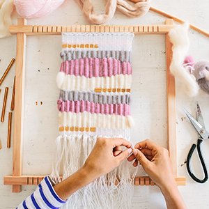 Hands weaving on a wooden loom with pink, grey, and white yarn, scissors and yarn spools nearby