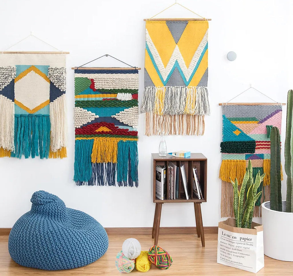 Four woven wall hangings displayed with a blue beanbag chair, yarn balls, a wooden side table with books, and potted plants.